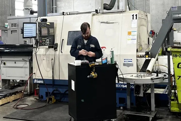 Technician working on a component in a CNC machining workshop, surrounded by industrial equipment including a large CNC machine, a work cart with tools, and a computer monitor displaying data